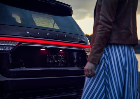 A person is shown near the rear of a 2024 Lincoln Aviator® SUV as the Lincoln Embrace illuminates the rear lights | Maguire's Lincoln in Palmyra PA